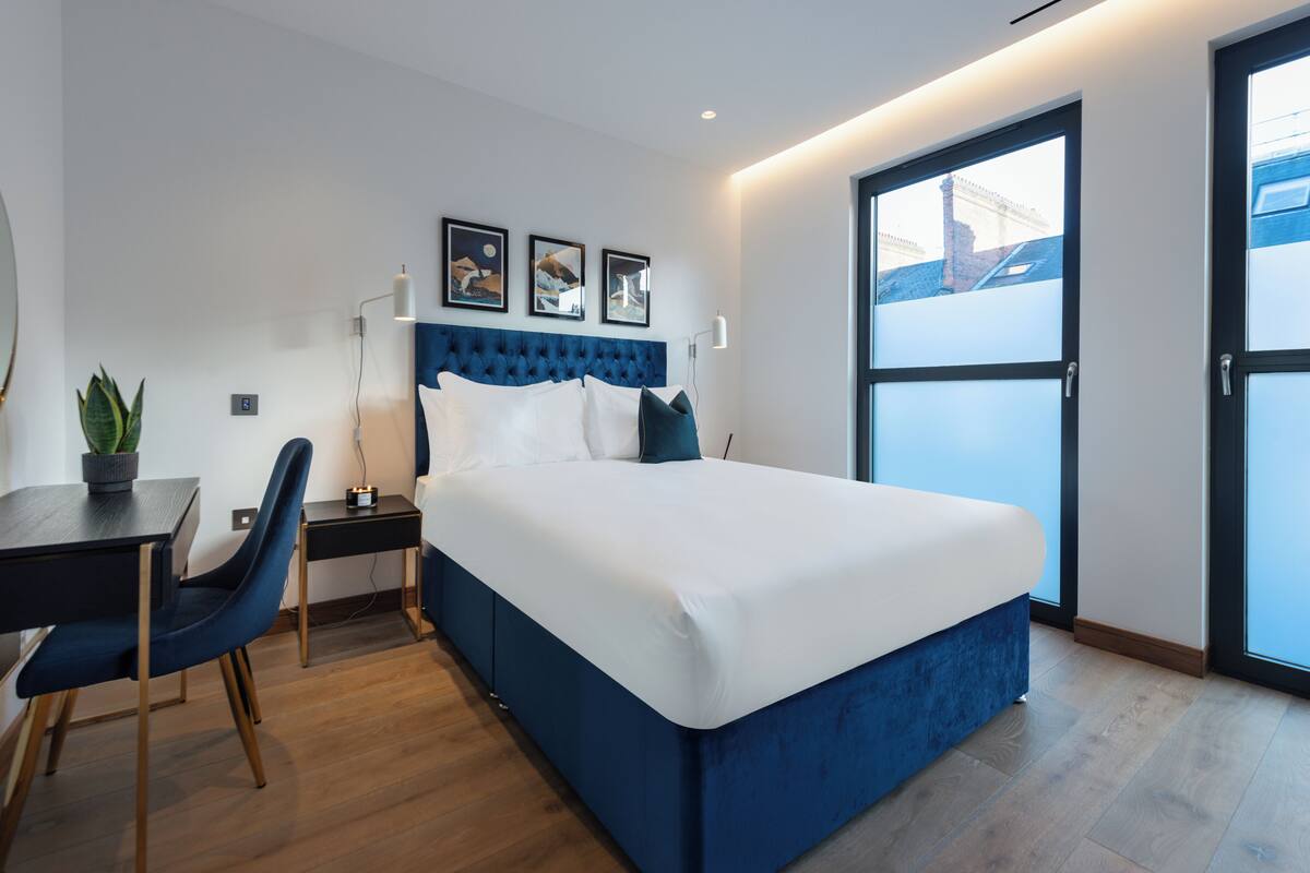 A modern hotel room with a large bed, a desk, a chair, a potted plant, and a view of the city through a large window.