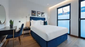 A modern hotel room with a large bed, a desk, a chair, a potted plant, and a view of the city through a large window.