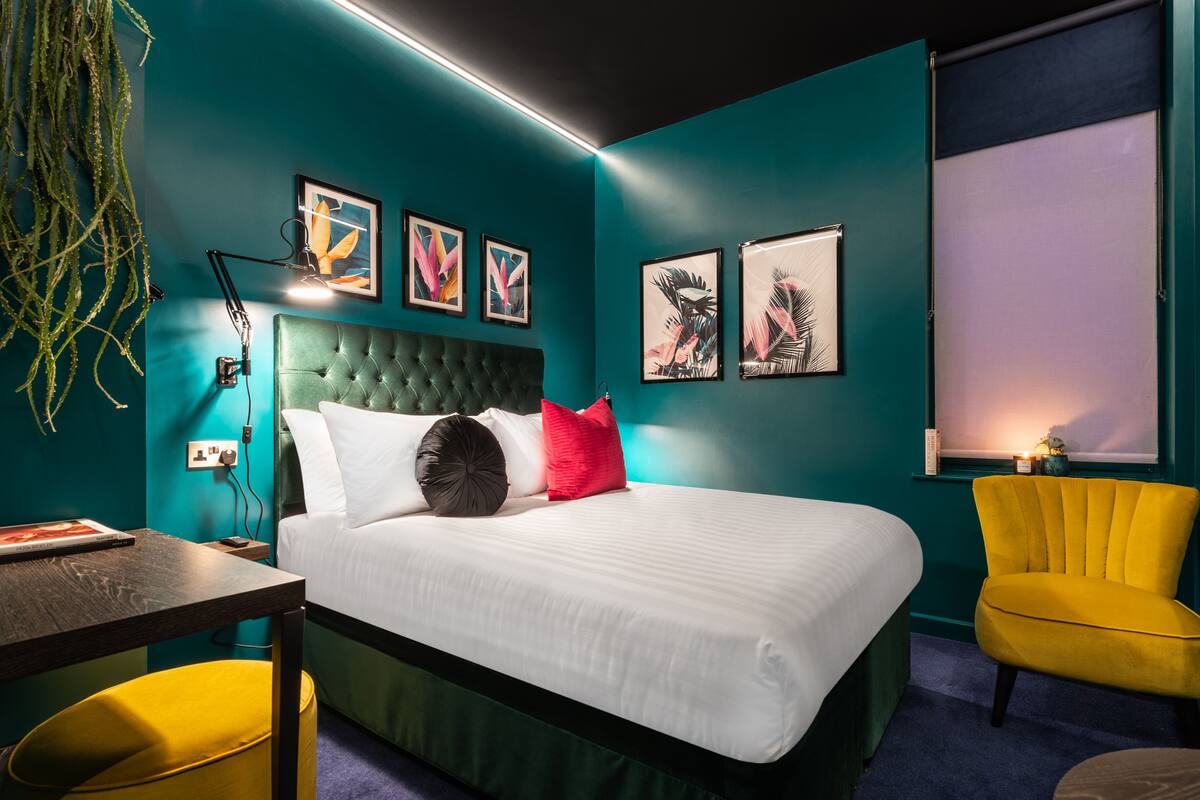 A modern bedroom with teal walls, a tufted headboard, a white bed, a yellow armchair, and artwork on the walls.