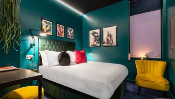 Un dormitorio moderno con paredes verde azulado, un cabecero capitonado, una cama blanca, un sillón amarillo y obras de arte en las paredes.