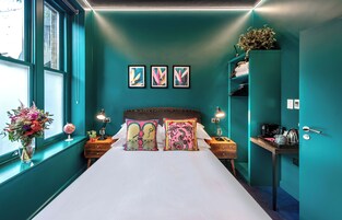 Ein schlafgemütliches Zimmer in Türkis mit einem großen Bett, zwei Nachttischen, einem Schminktisch und einem Kleiderschrank, sowie einem Fenster mit Blick.