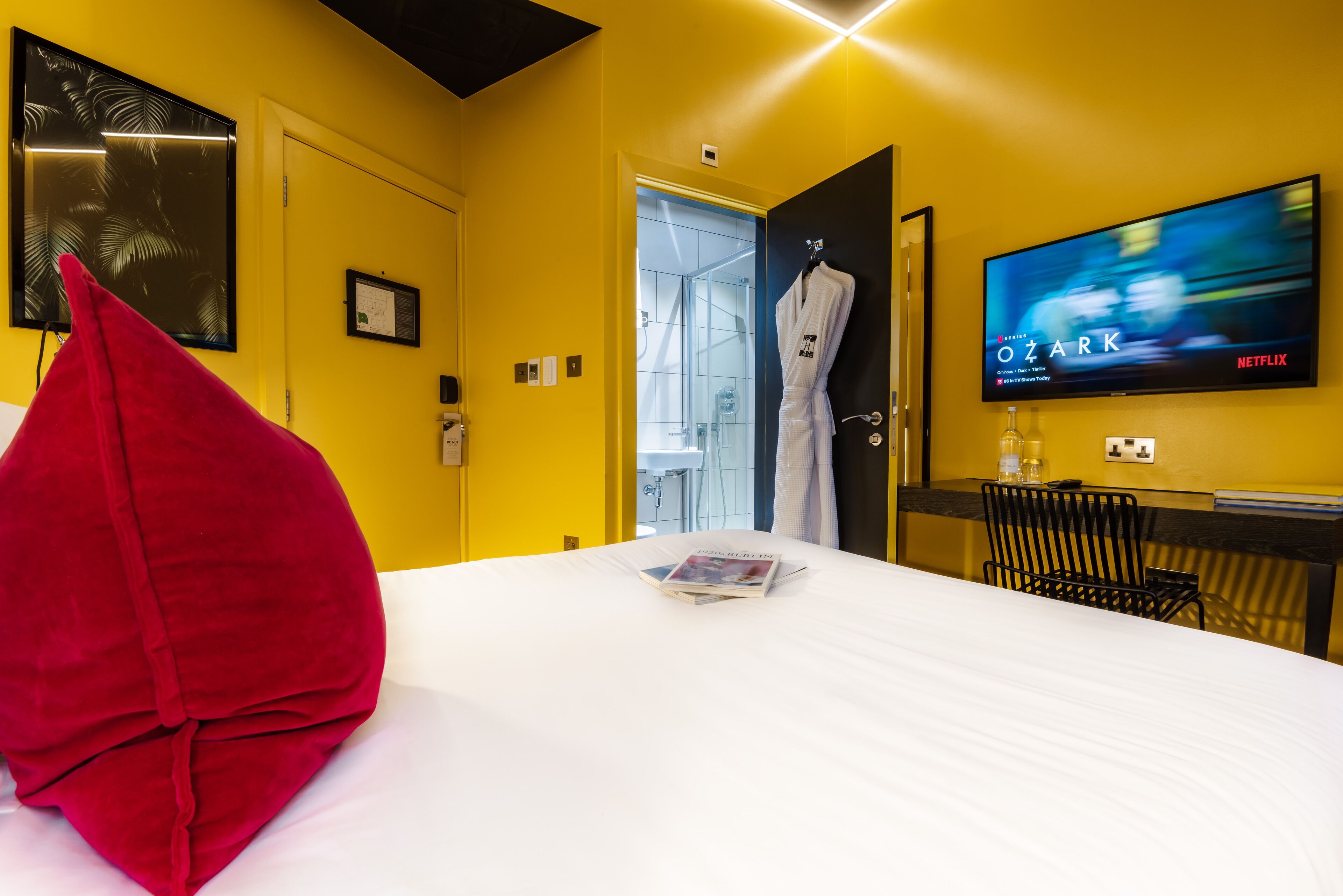 A hotel room with a bed, a red pillow, a TV displaying 'Ozark', and a robe hanging on the door.