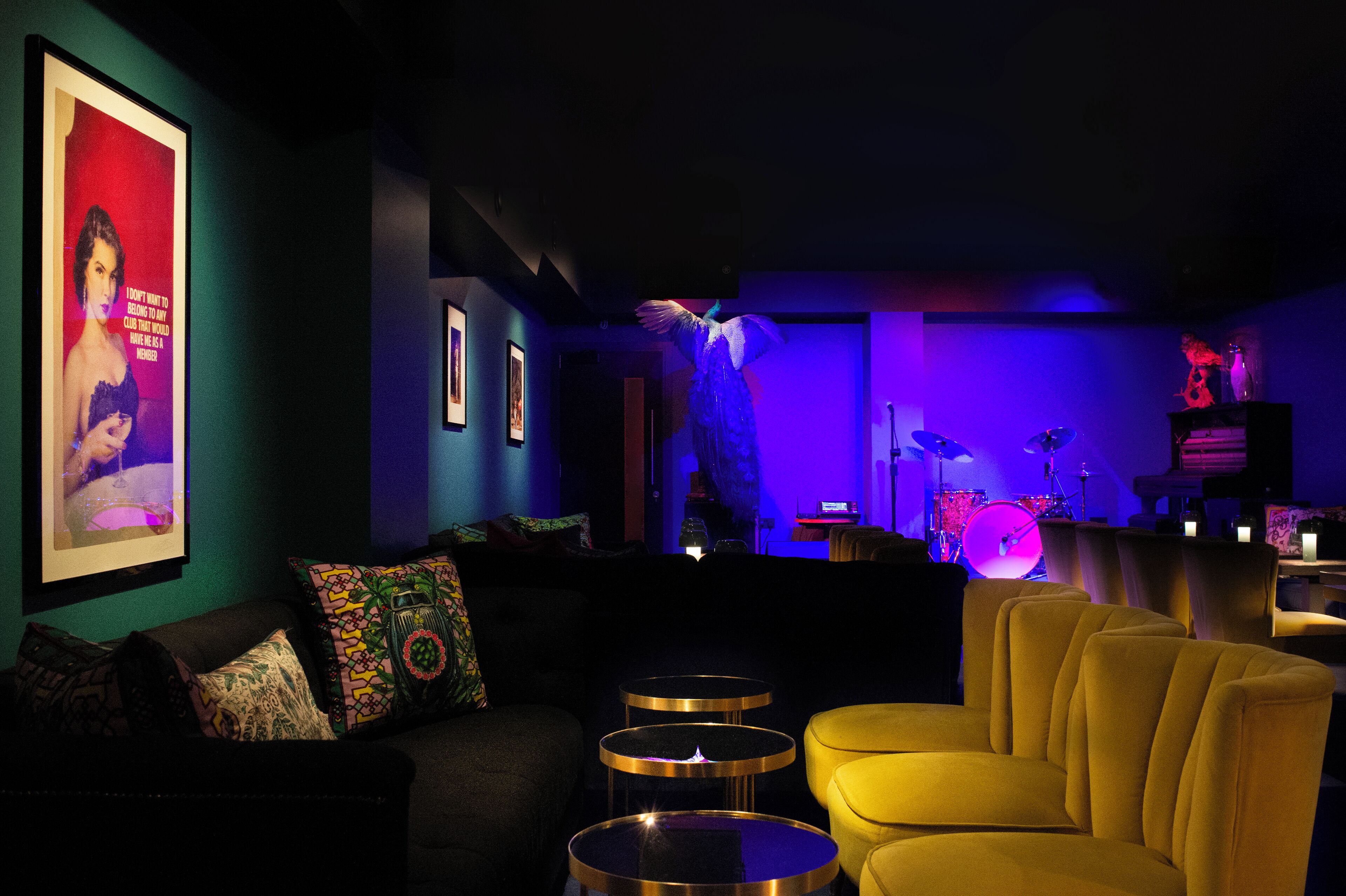 Una habitación con poca luz, un sillón amarillo, un sofá negro con cojines estampados y un cartel en la pared.