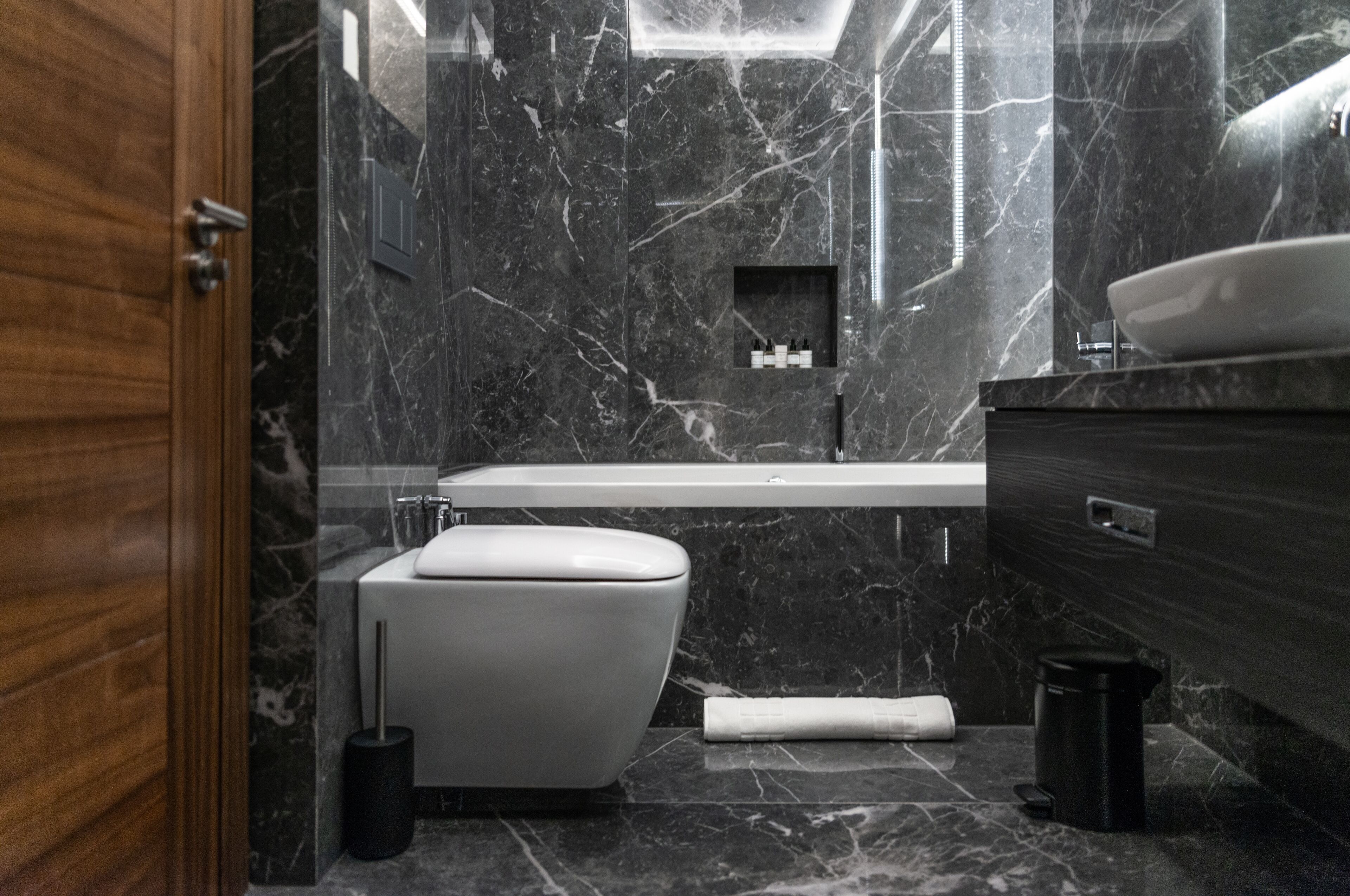 A modern bathroom with a marble wall, a white toilet, a bathtub, and a wooden vanity.