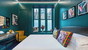 A modern bedroom with a large bed, a desk with a chair, a window with a view of buildings, and artwork on the walls.