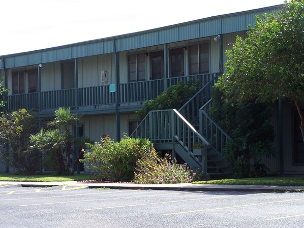 A multi-story building with a balcony on the second floor, a parking lot in front, and some greenery around. - Mustang Bungalow 4MI (Port Aransas)
