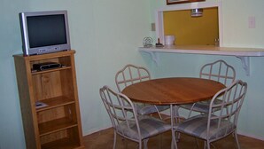 Un comedor con una mesa redonda de madera, cuatro sillas de metal, un televisor sobre un soporte de madera, un reloj de pared y una lámpara colgante.
