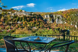 A table with a plate of food and glasses, set against a backdrop of a mountainous landscape with buildings on a hill. - En Chora Vezitsa (Zagori)