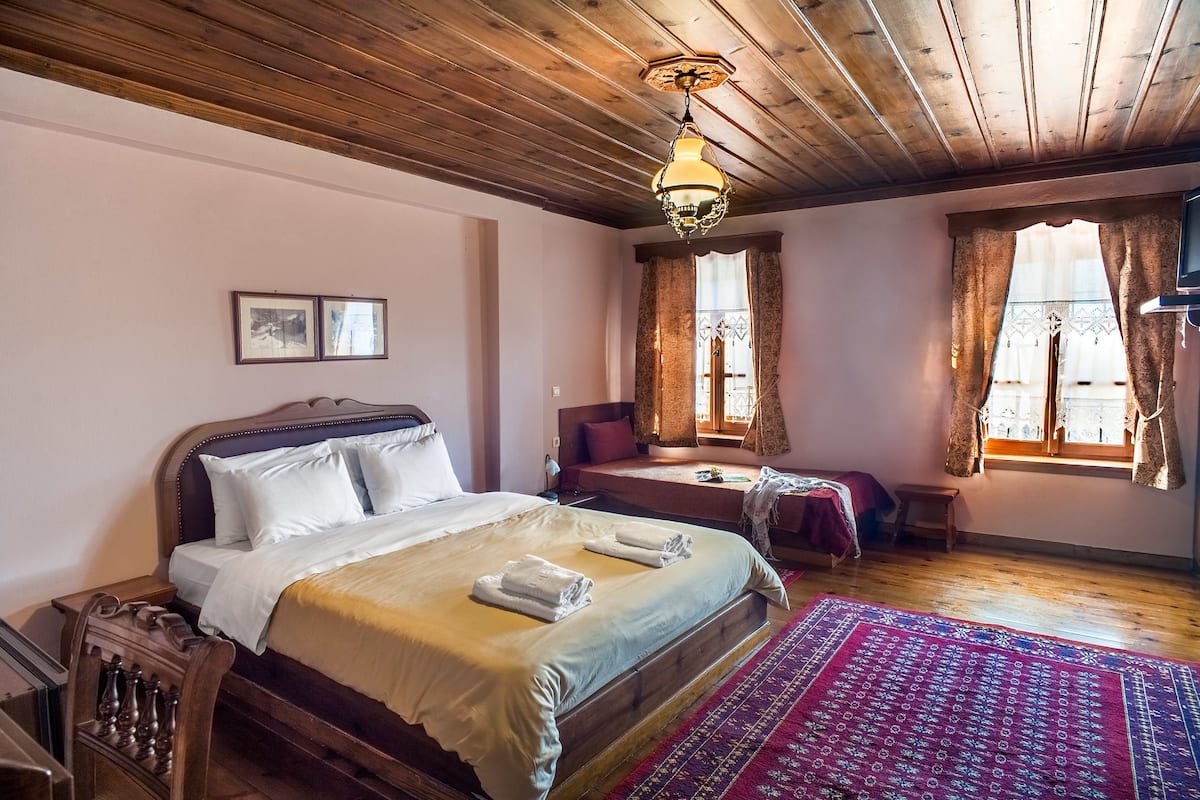 A bedroom with a wooden ceiling, a large bed with white linens, a red patterned rug, and two windows with curtains.