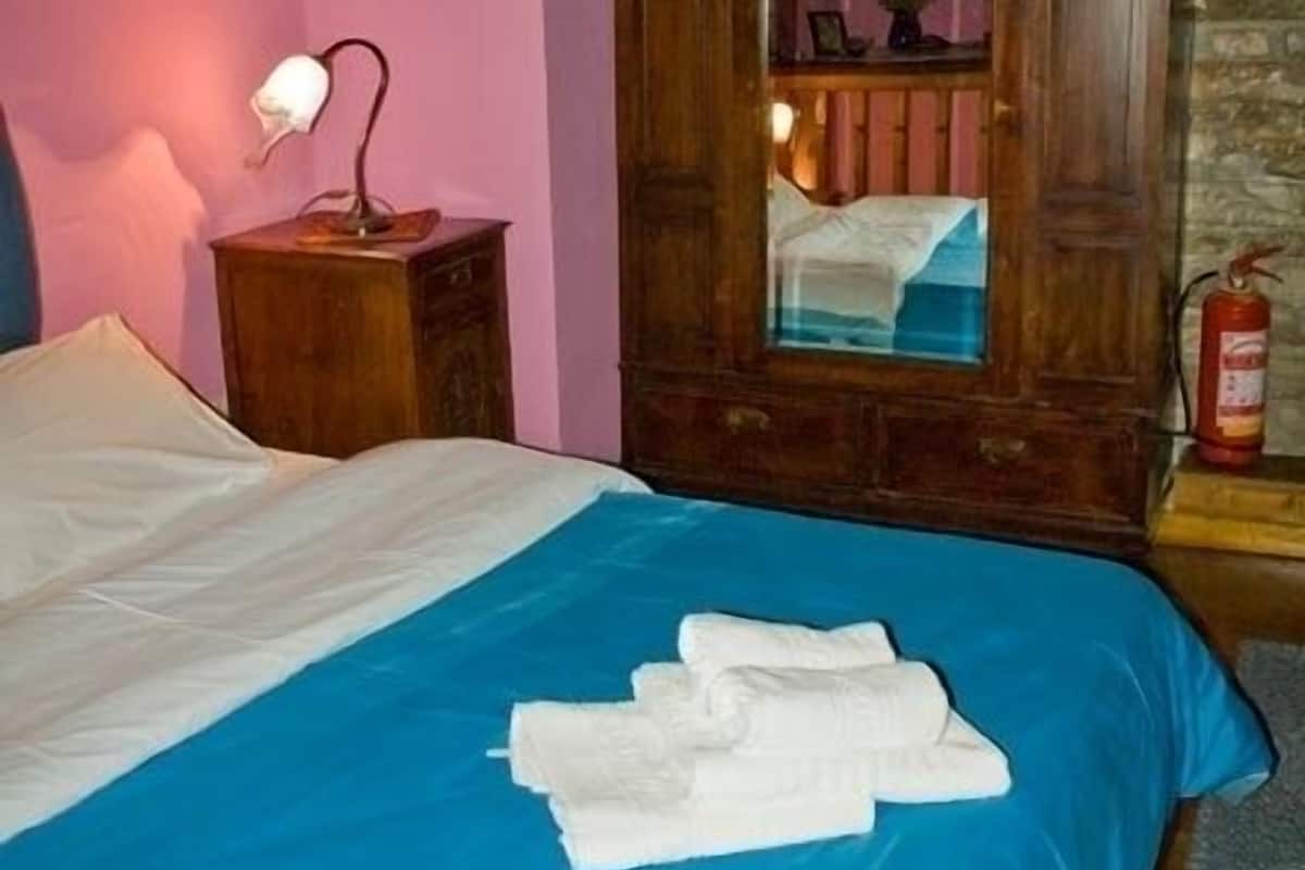 A hotel room with a bed, a wooden wardrobe, a nightstand with a lamp, and a fire extinguisher.