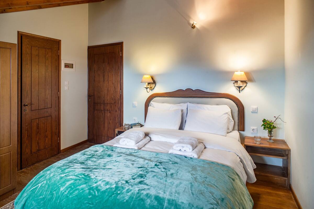 A bedroom with a large bed, wooden wardrobe, two bedside lamps, and a nightstand with a glass.