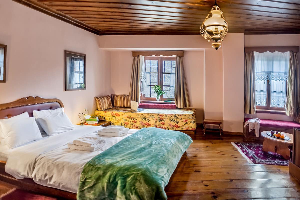 A bedroom with a large bed, a desk with a laptop, a window with curtains, and a wooden ceiling.
