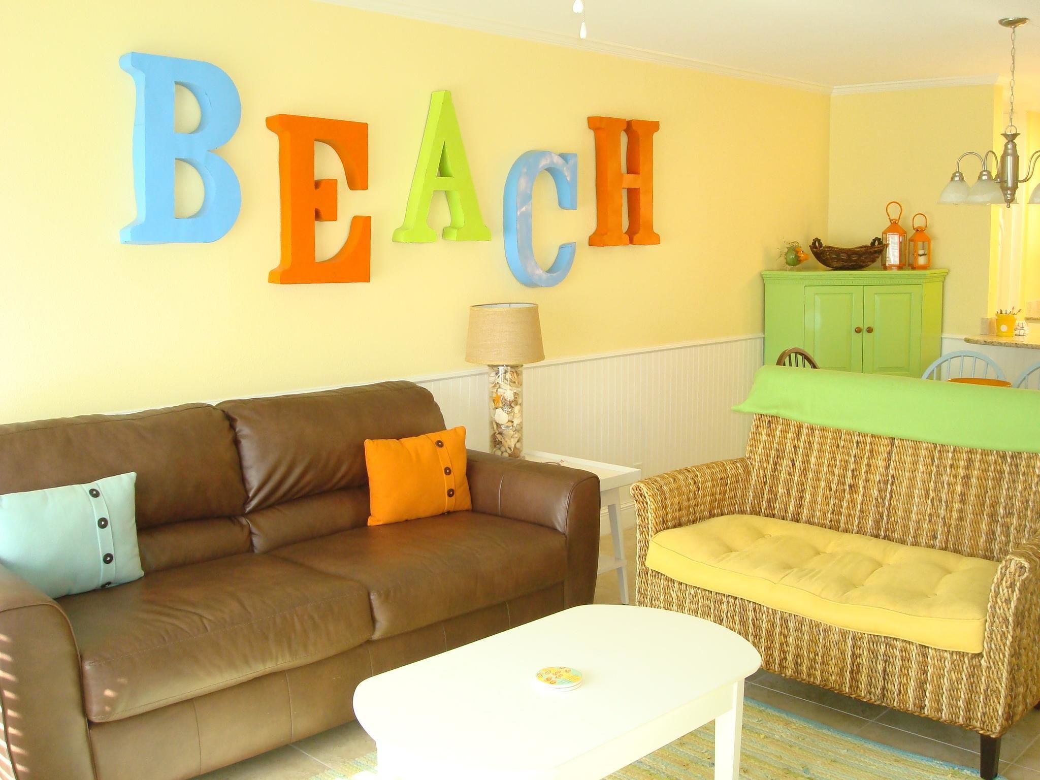 A living room with a brown leather sofa, a white coffee table, and a wicker armchair. A wall-mounted word "BEACH" is visible.