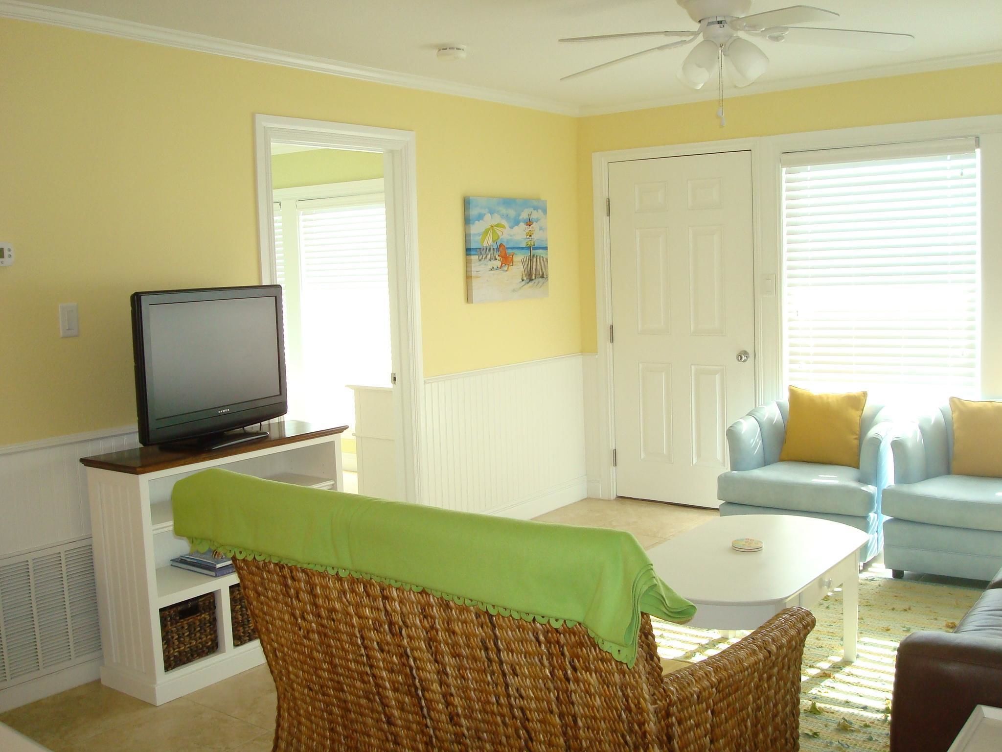 A living room with a television on a stand, a wicker chair, a white coffee table, a blue sofa, and a door with a painting above it.