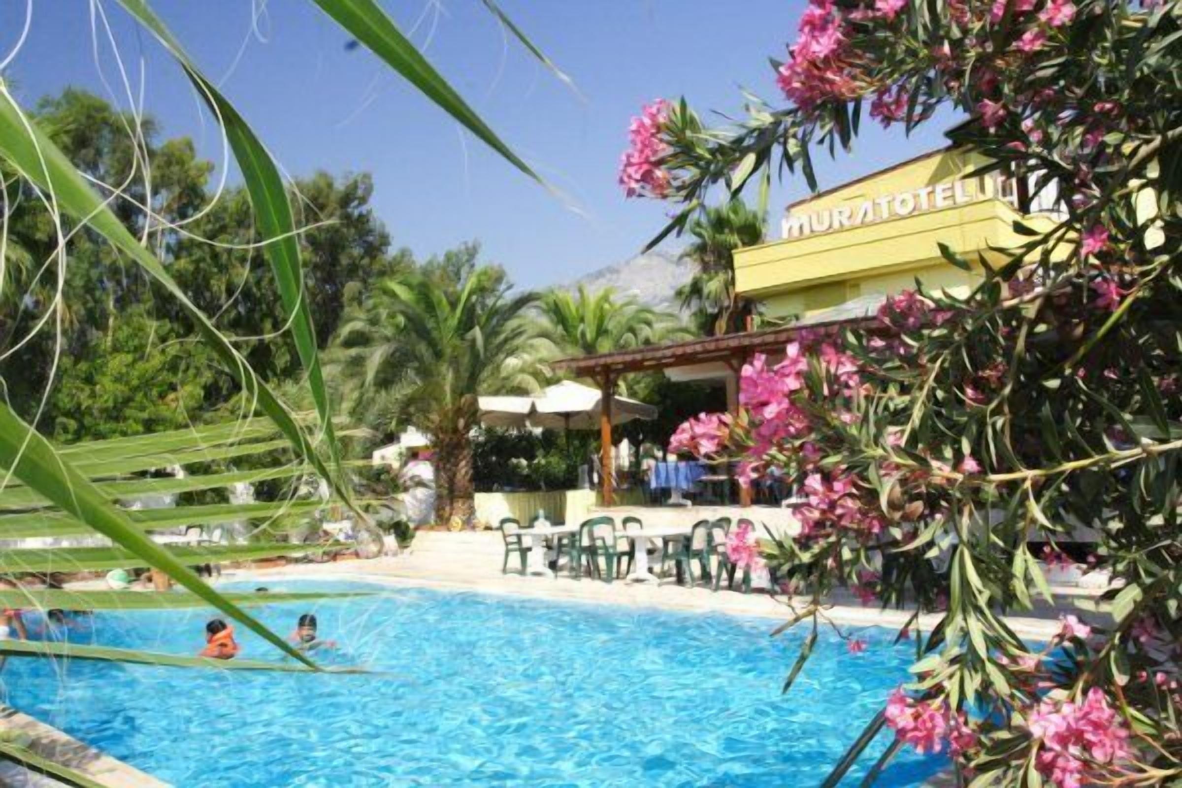 A swimming pool area with a building labeled "MURATOTEL" in the background, surrounded by greenery and palm trees.