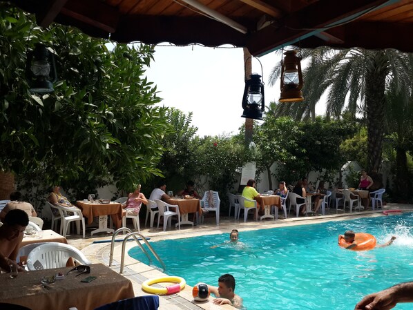 An outdoor pool area with tables and chairs, surrounded by greenery. - Murat Hotel (Kemer)