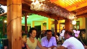 A wooden bar area with people seated at tables, a green-lit background, and a hanging light fixture. - Murat Hotel (Kemer)