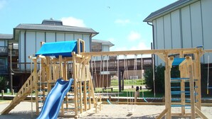 A playground with a slide and climbing structures, surrounded by residential buildings. - Sunrise View 303BH (Port Aransas)