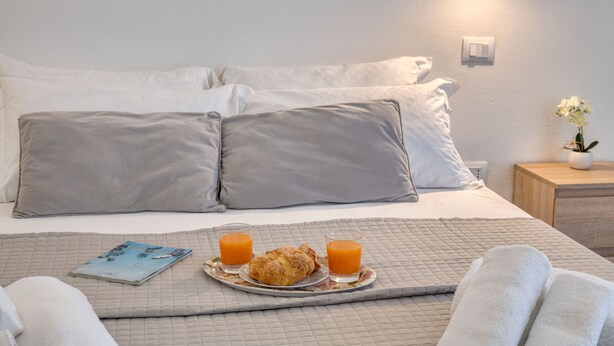 Una cama bien hecha con almohadas, una bandeja con cruasanes y jugo de naranja, y una mesita de noche con una planta.