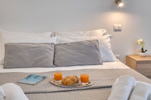 Una cama bien hecha con almohadas, una bandeja con cruasanes y jugo de naranja, y una mesita de noche con una planta.
