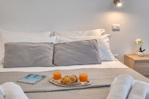 Una cama bien hecha con almohadas, una bandeja con cruasanes y jugo de naranja, y una mesita de noche con una planta.
