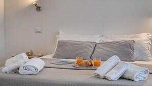 Una cama bien hecha con almohadas, una mesita de noche con un vaso de jugo de naranja, una toalla doblada y una bandeja con cruasanes y un libro.