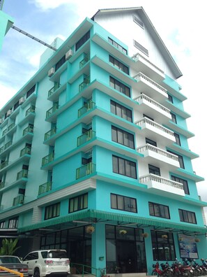 Un hotel de varios pisos con fachada azul verdosa y blanca, balcones en cada planta y un área de entrada cubierta.