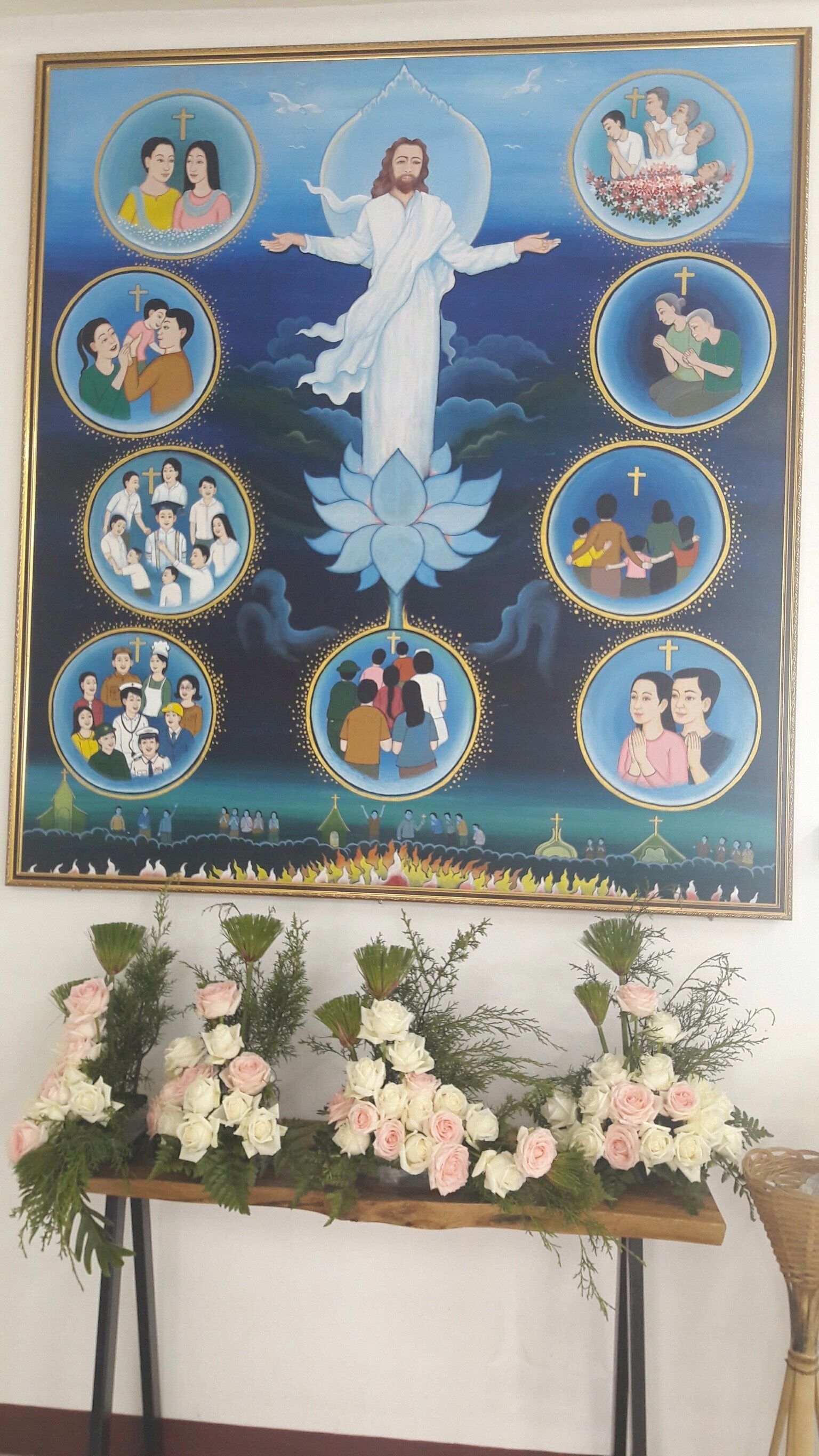 Una pintura religiosa con una figura central rodeada de escenas más pequeñas, exhibida sobre un arreglo floral.