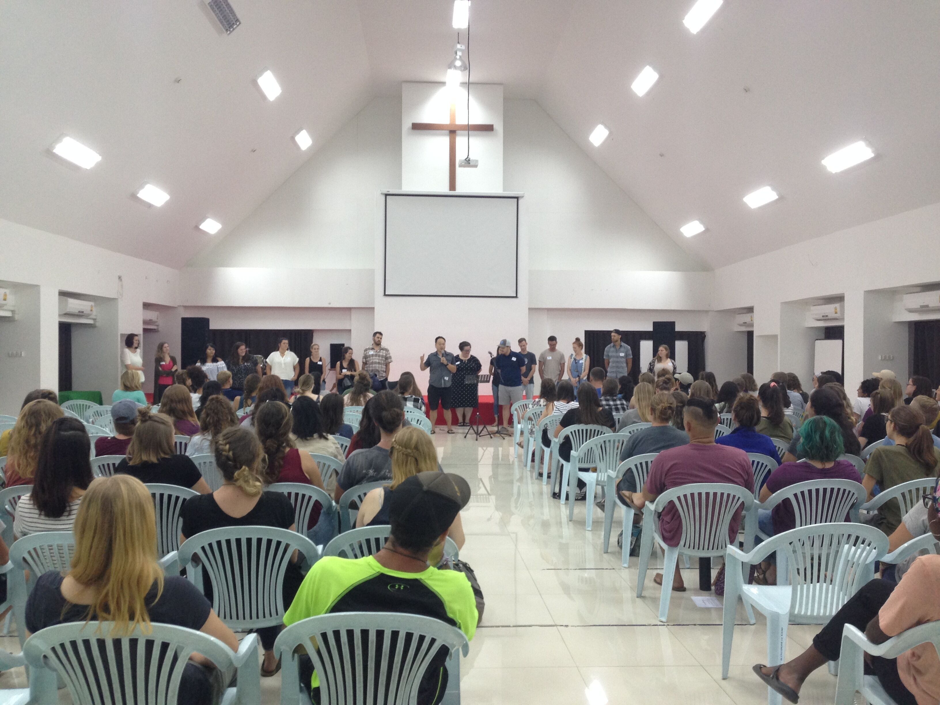 A large hall with rows of white plastic chairs, a stage with speakers, and a cross on the wall.