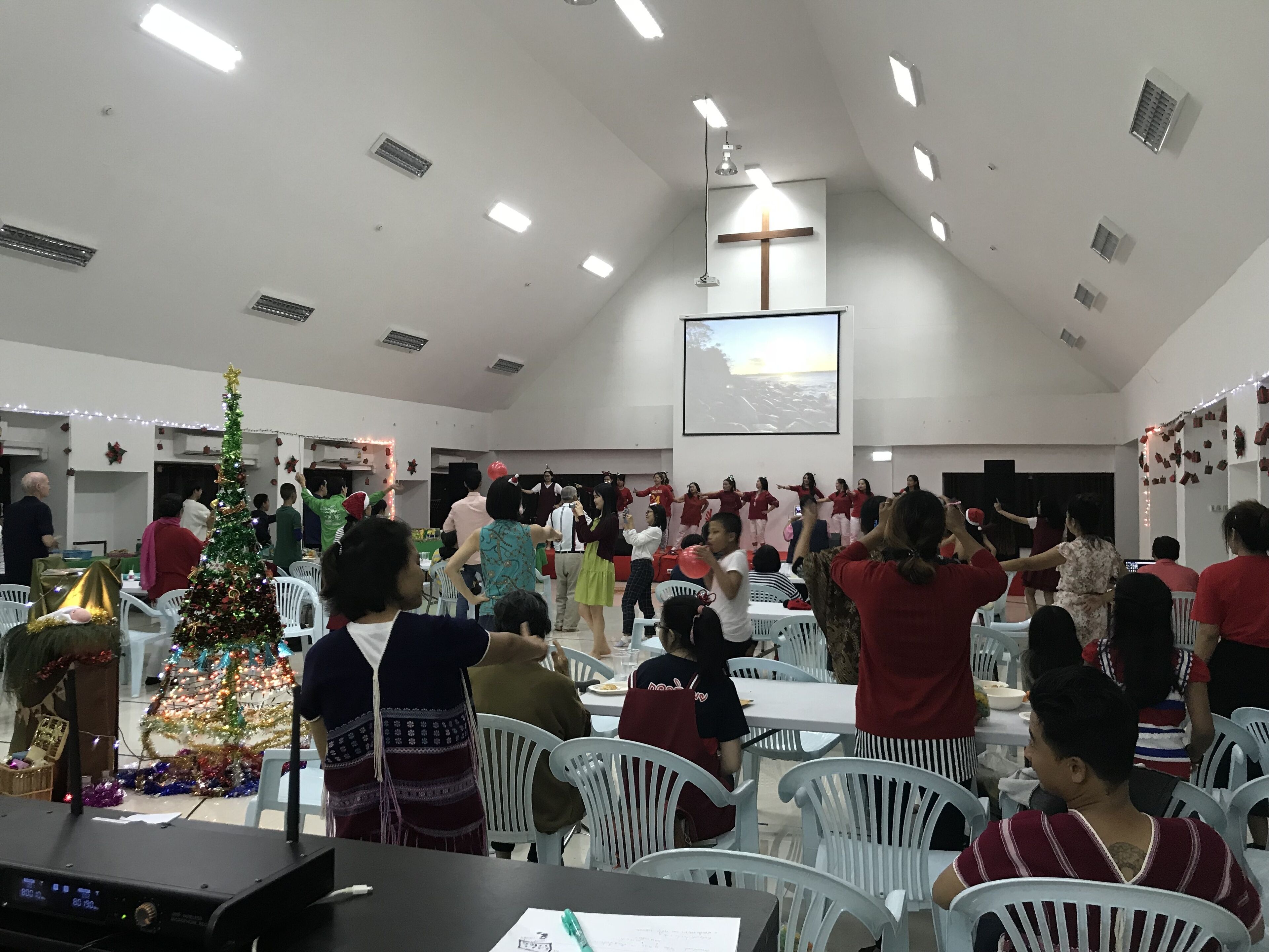 Um salão amplo com paredes brancas, uma grande cruz na parede e um pinheiro de Natal.