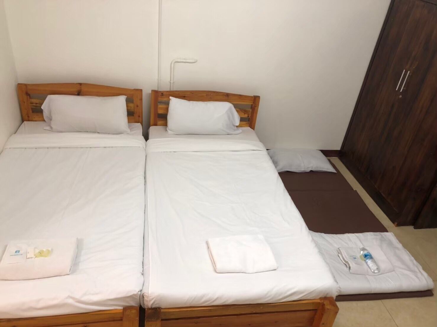 A room with two wooden beds, white bedding, a brown mat, a wooden wardrobe, and a white towel on each bed.