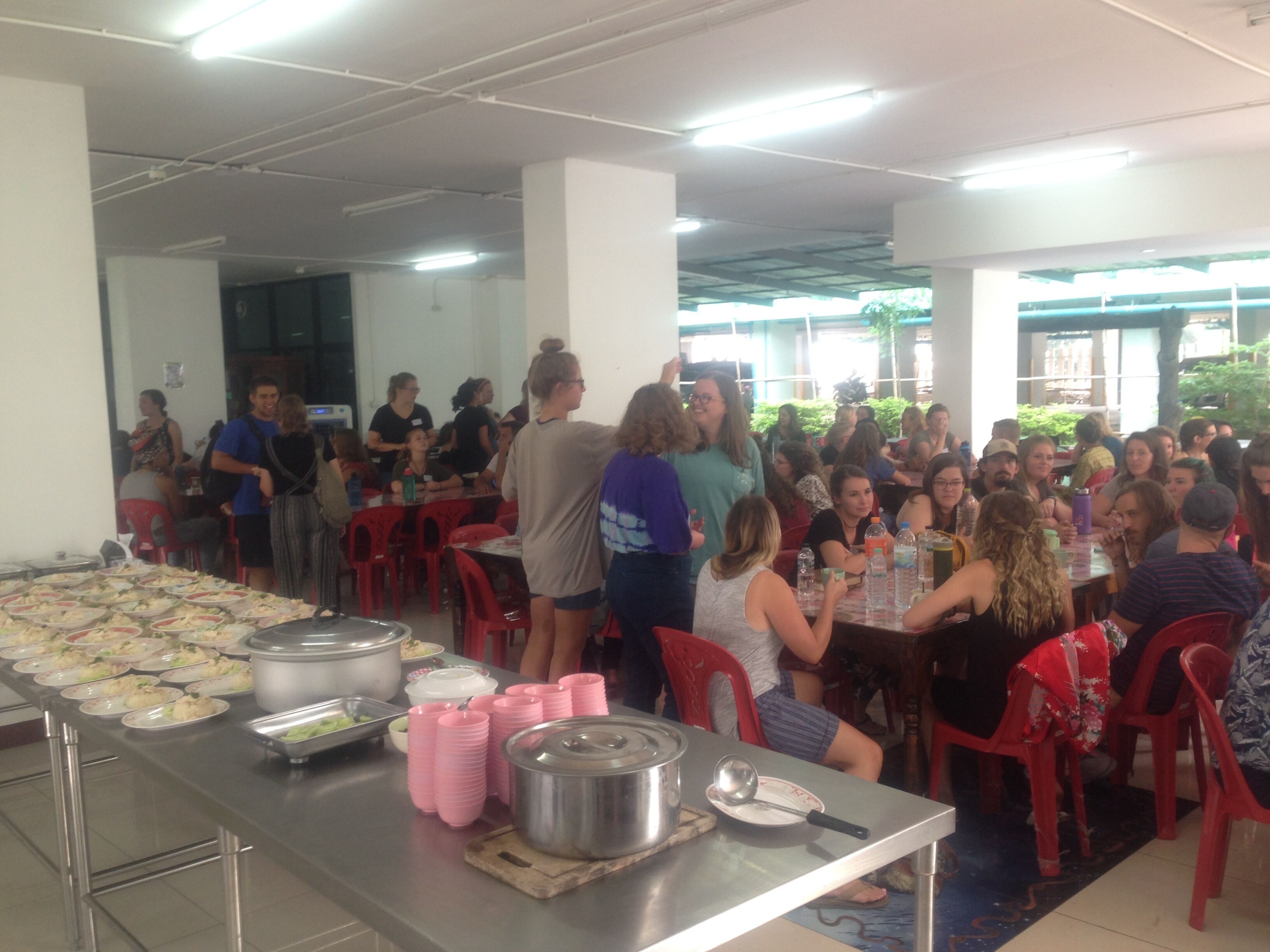 A cafeteria with tables and chairs, food trays, and serving utensils.
