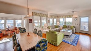 A spacious living room with a wooden dining table, multiple seating options, and large windows offering a view. - Bonnie Doon 2 Bedroom Holiday Home By Bald Head Island (Bald Head Island)