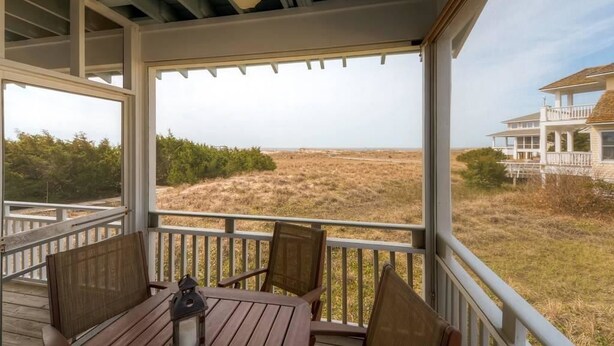 A covered outdoor area with a table and chairs, overlooking a grassy landscape and a distant building. - Bonnie Doon 2 Bedroom Holiday Home By Bald Head Island (Bald Head Island)
