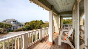 Een houten terras met witte leuningen, een overdekte veranda en uitzicht op de oceaan en omliggende huizen.