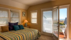 A bedroom with a bed, a nightstand, a lamp, and a view of the balcony. - Blue Heron 3 Bedroom Holiday Home By Bald Head Island (Bald Head Island)