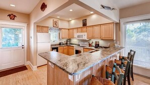 A kitchen with wooden cabinets, a marble countertop, and a built-in island with bar stools. - Blue Heron 3 Bedroom Holiday Home By Bald Head Island (Bald Head Island)
