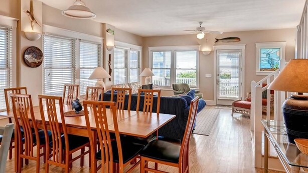 A spacious living area with a dining table, chairs, and a sofa. There are large windows with blinds, a ceiling fan, and wall-mounted lamps. - Blue Heron 3 Bedroom Holiday Home By Bald Head Island (Bald Head Island)
