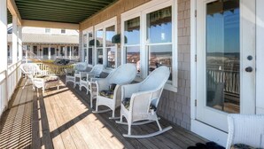 A covered porch with white wicker rocking chairs, a wooden deck, and a view of a building in the background. - Bogan s on the Beach 4 Bedroom Holiday Home By Bald Head Island (Bald Head Island)