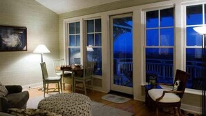 A cozy room with a dining table, chairs, a sofa, and a window with a view of a balcony. - Cape Watch Cottage 4 Bedroom Holiday Home By Bald Head Island (Bald Head Island)