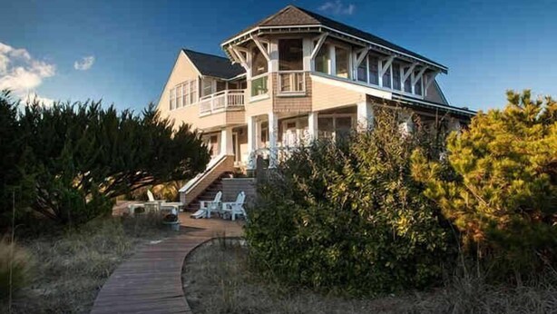 A large, two-story house with a prominent balcony and a wooden deck leading to the entrance. - Cape Watch Cottage 4 Bedroom Holiday Home By Bald Head Island (Bald Head Island)