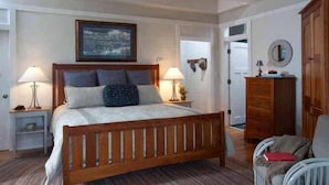 A bedroom with a wooden bed, two bedside lamps, a framed picture on the wall, a wardrobe, and a chair. - Cape Watch Cottage 4 Bedroom Holiday Home By Bald Head Island (Bald Head Island)