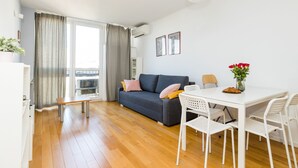Un salon moderne avec un canapé gris, des chaises blanches, une petite table et une fenêtre avec des rideaux.
