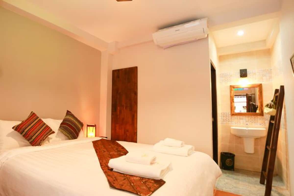 Une chambre d’hôtel comprenant un lit, des oreillers, une table de chevet, un appareil de climatisation, une salle de bain avec un lavabo et un miroir, et une porte en bois.