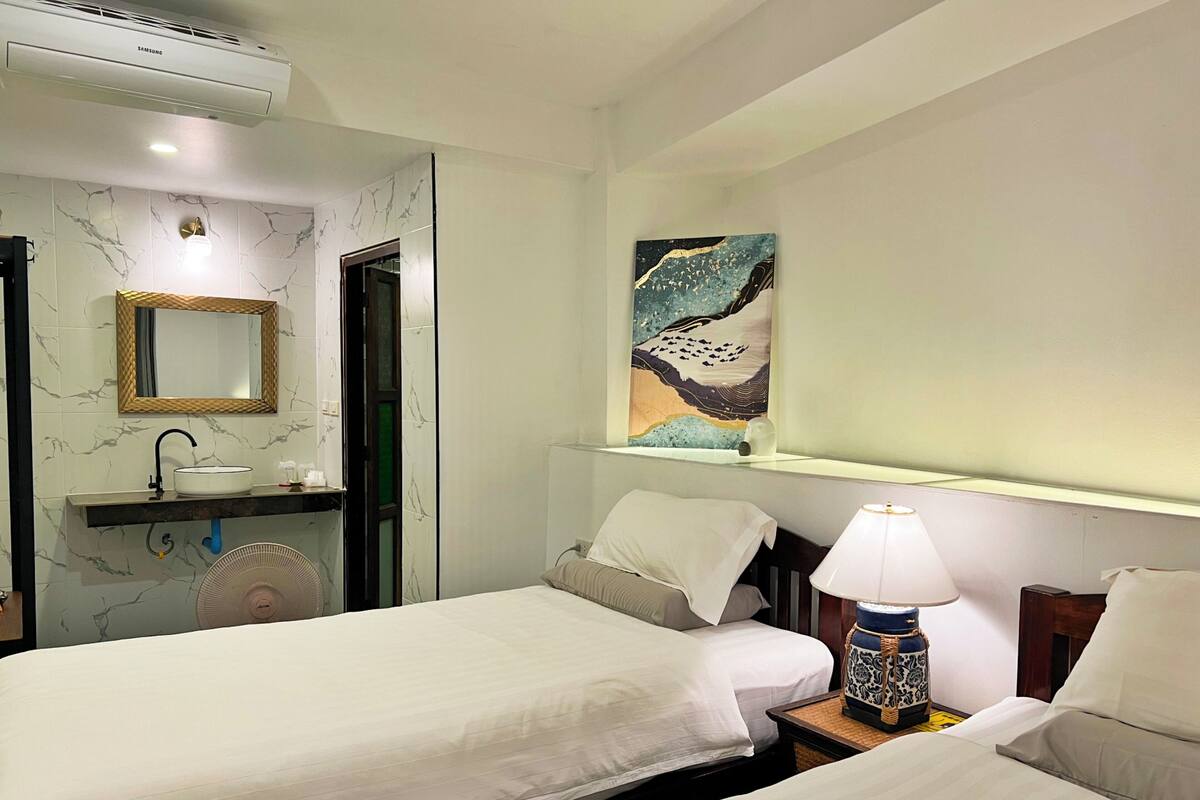 Une chambre d’hôtel avec deux lits, une salle de bain comprenant un lavabo, un miroir et un ventilateur, ainsi qu’une étagère fixée au mur où est posé un objet décoratif.