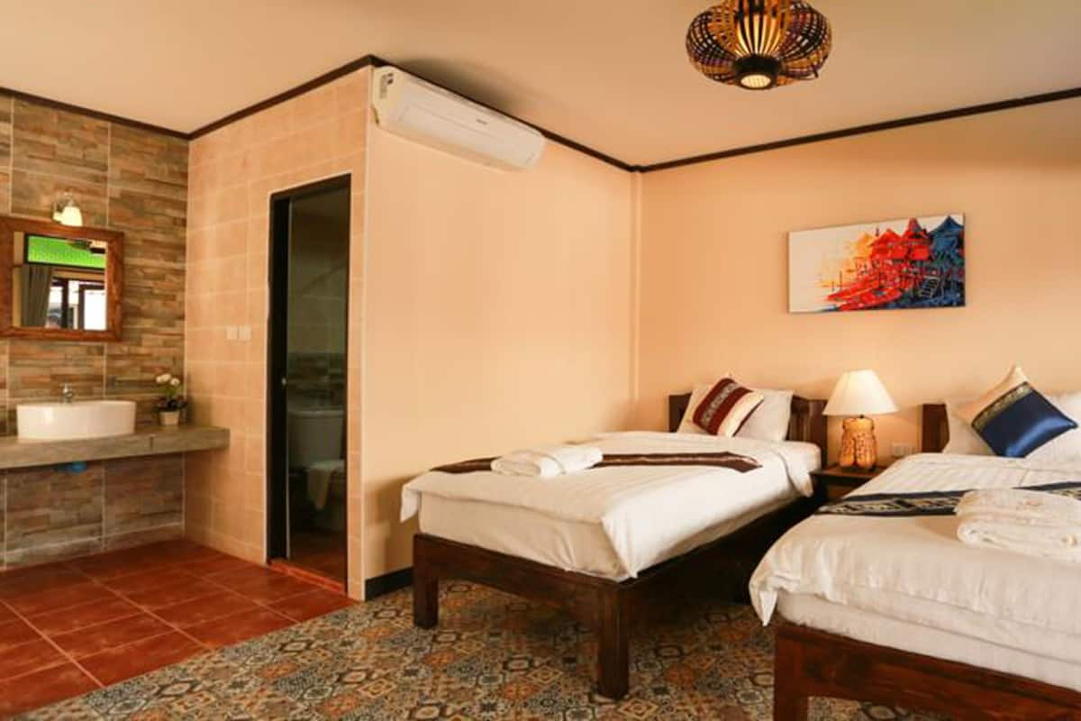 Une chambre d’hôtel avec deux lits, une salle de bain dotée d’un lavabo et d’un miroir, un tapis à motifs et un tableau représentant un paysage urbain accroché au mur.