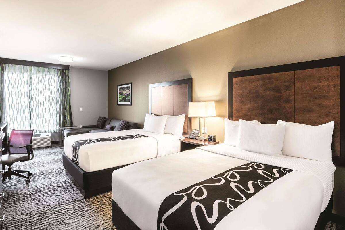 Habitación de hotel con dos camas, un escritorio, una silla, una lámpara y un cuadro en la pared.