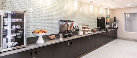 Een moderne keukenhoek met een lange toonbank, uitgerust met een koffieapparaat, een broodvitrine en een schaal fruit.
