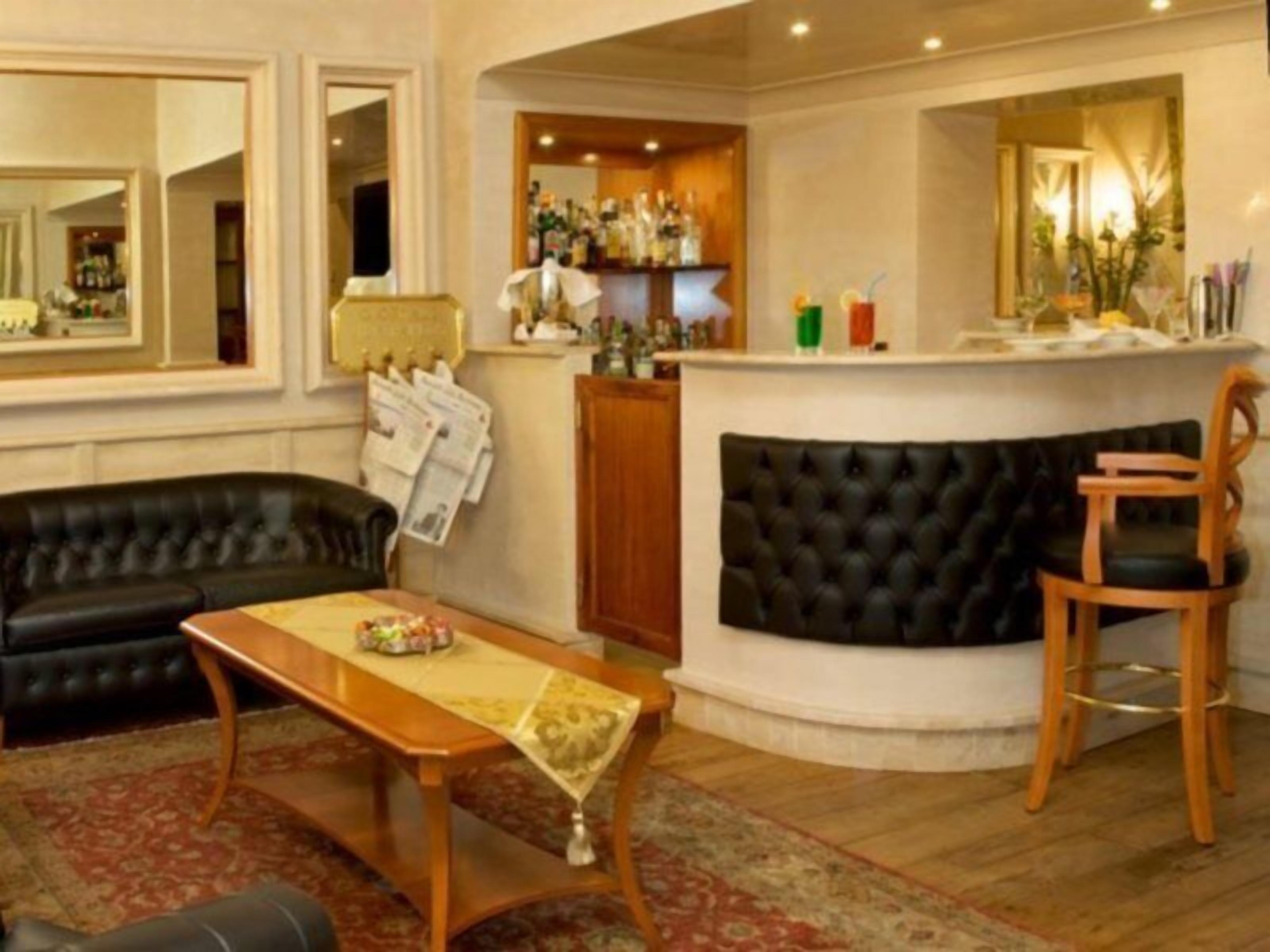 A room with a black leather sofa, a wooden table with a decorative tablecloth, a bar area with a mirror and various bottles, and a wooden chair.