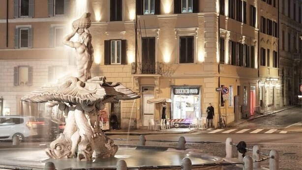 A classical fountain with a statue in the center, surrounded by a paved area with bollards. In the background, there are multi-story buildings with balconies and windows. - Barocco (Rome)